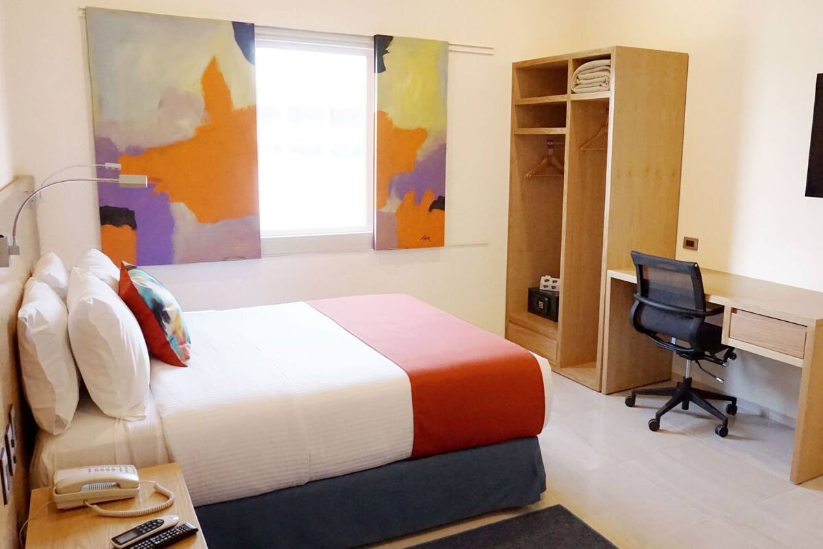 Une chambre d’hôtel comprenant un lit, une table de chevet, un téléphone, des télécommandes, un bureau avec une chaise, une armoire et des tableaux abstraits accrochés aux murs.
