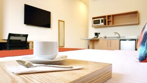 Une chambre d’hôtel avec un lit, une table de chevet sur laquelle se trouvent une tasse, une cuillère et un livre. Un téléviseur est fixé au mur et il y a un four à micro-ondes dans le coin cuisine.