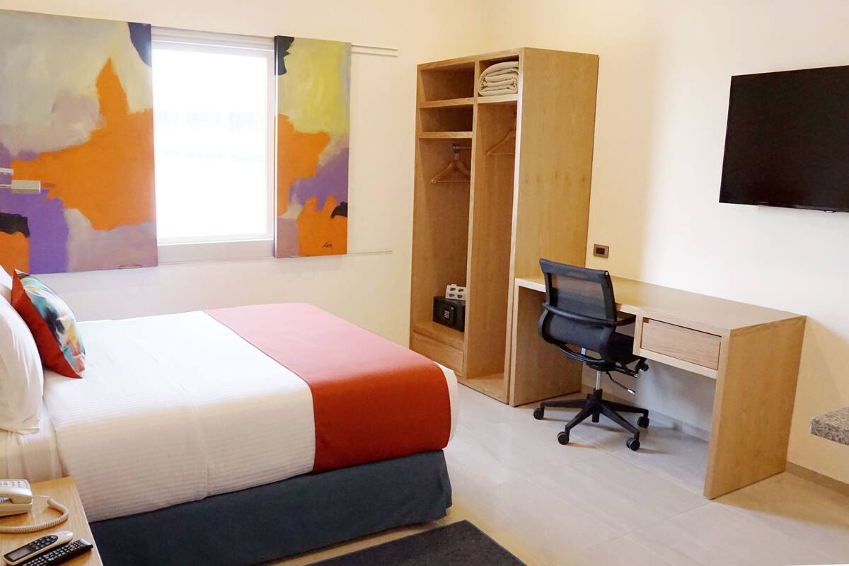Une chambre d’hôtel équipée d’un lit, d’une table de chevet, d’un téléphone et d’un bureau avec une chaise. On y trouve également une armoire et un téléviseur fixé au mur.