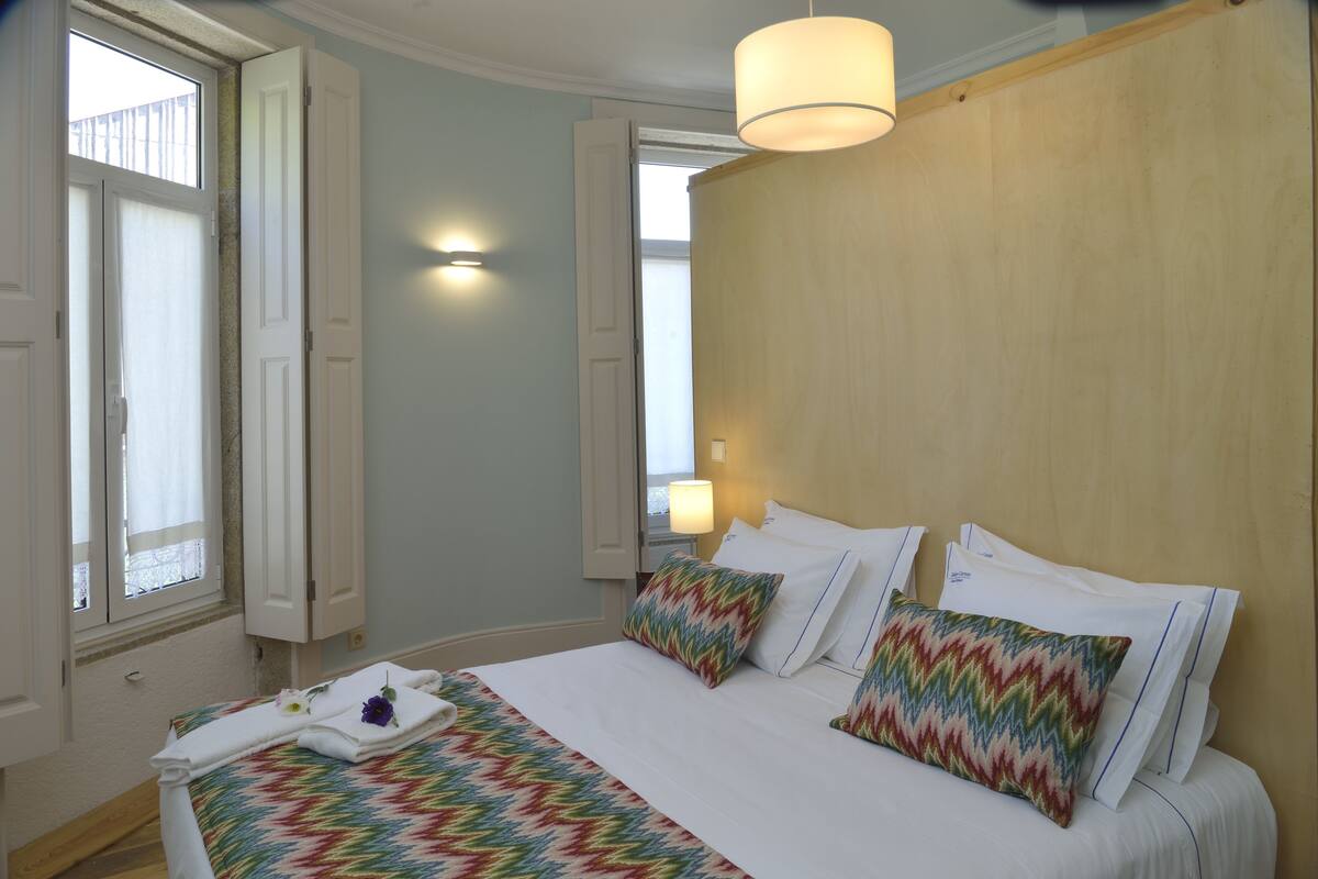 Ein Hotelzimmer mit Bett, Vorhängen am Fenster, einer Wandlampe und einem Holzkopfteil.