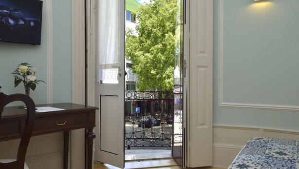 Une pièce avec un bureau en bois, une chaise, un lit avec une literie à motifs, un téléviseur fixé au mur et une porte donnant sur une rue bordée d’arbres et où sont garées des voitures.