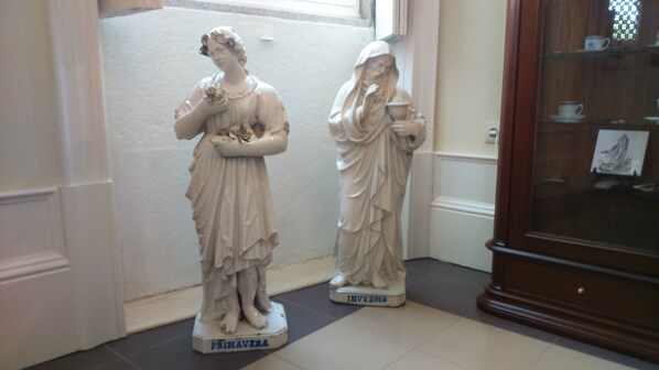 Dos estatuas blancas de mármol que representan a mujeres, sobre pedestales con las etiquetas 'PRIMAVZRA' e 'INVERNO', en una habitación con suelo de baldosas y un armario de madera con puertas de cristal.