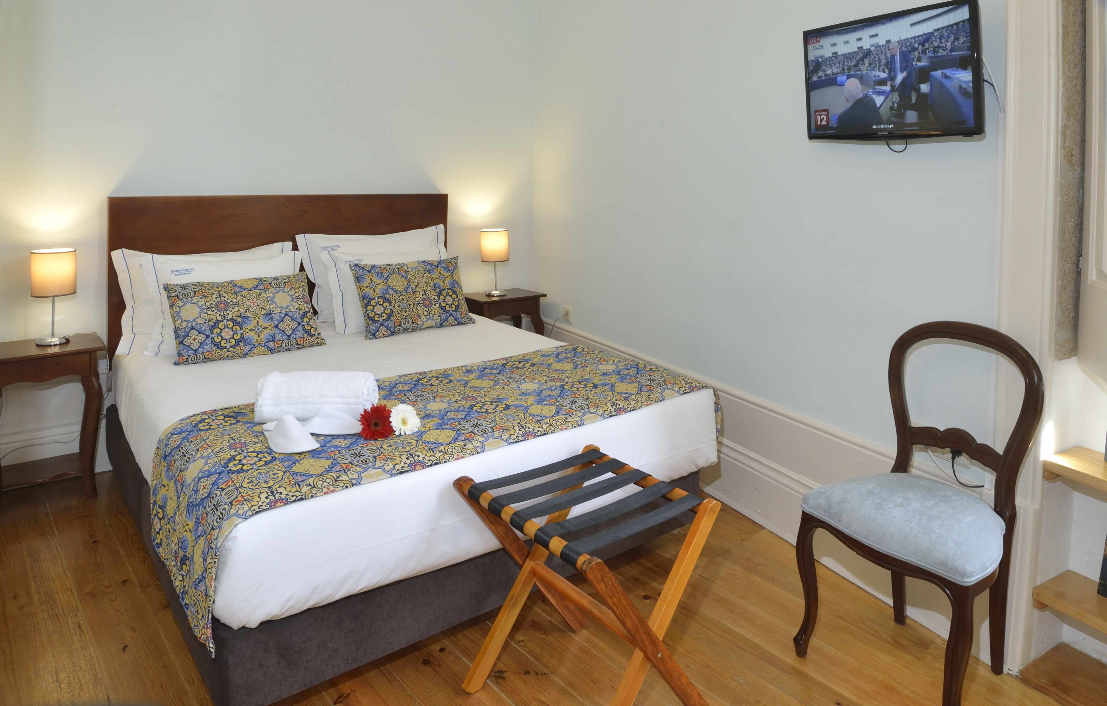 A neatly made bed with patterned bedding, a wooden chair, and a television mounted on the wall.