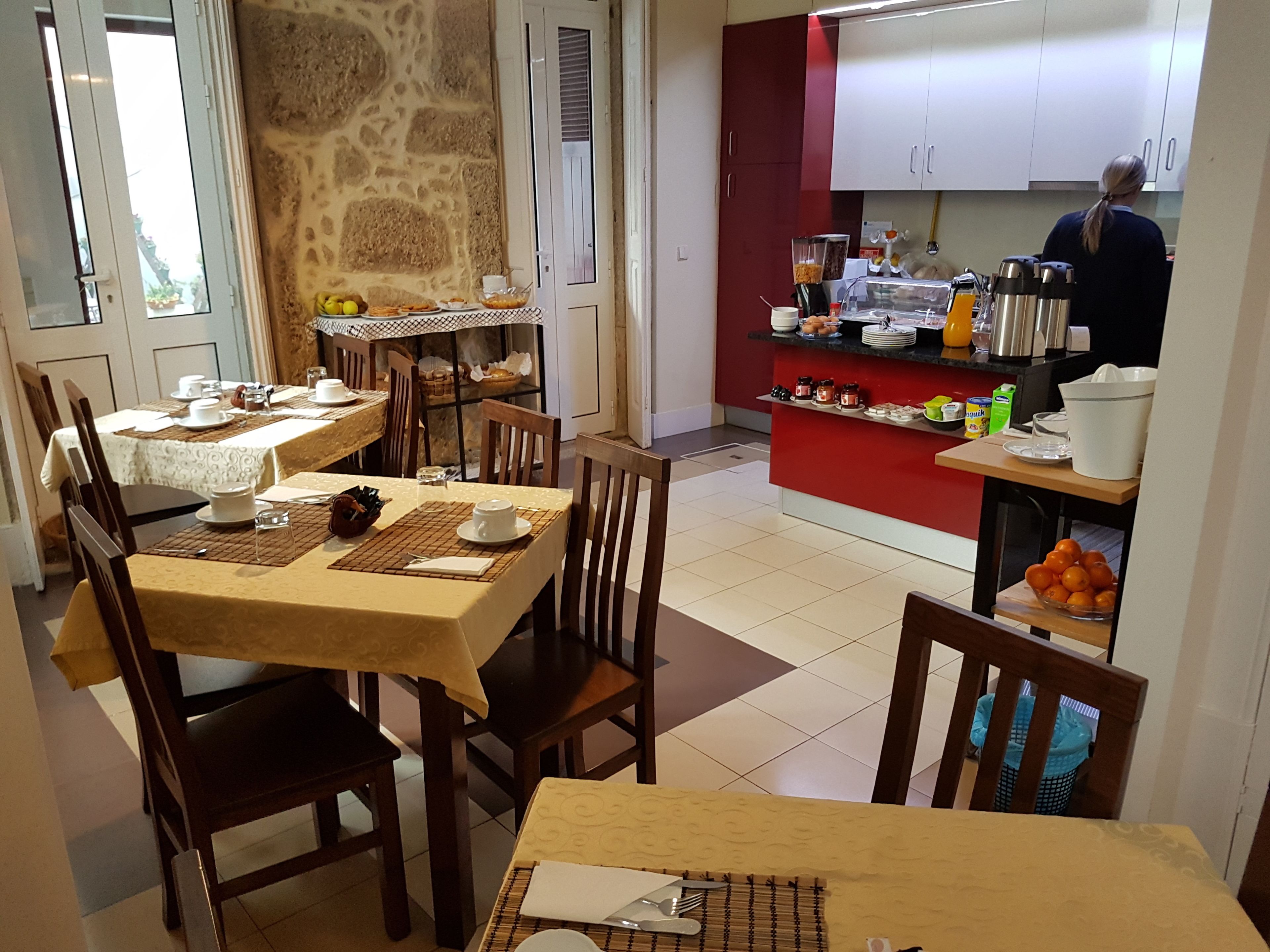 Un coin repas avec des tables préparées pour le petit-déjeuner, un espace cuisine équipé de divers appareils et contenant des provisions, et un mur en pierre.