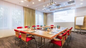 Sala de conferências com uma mesa comprida, cadeiras vermelhas e uma tela de projetor exibindo informações do hotel.