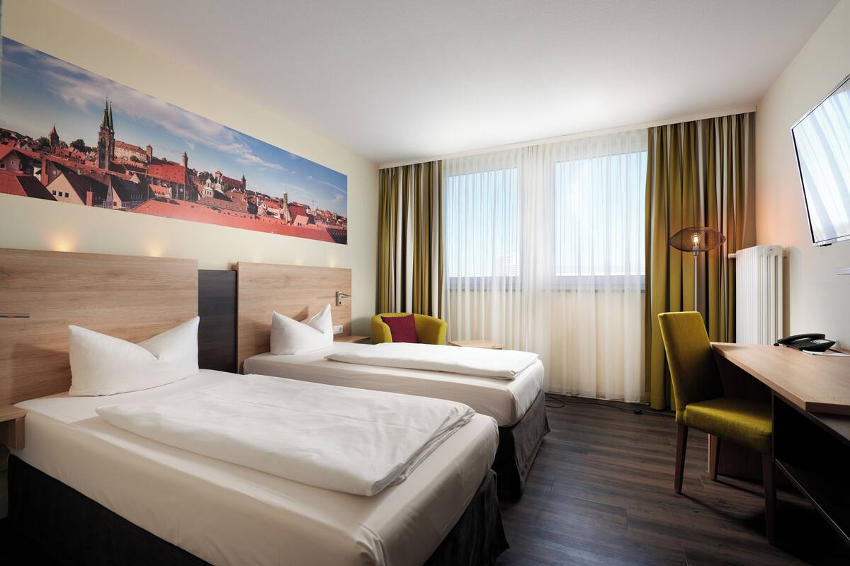A hotel room with two beds, a desk, a chair, a lamp, and a large window with curtains.