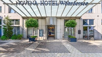 Exterior del Hotel Novina Wörthersee con una entrada moderna de vidrio, dos árboles en macetas y un aparcamiento para bicicletas.