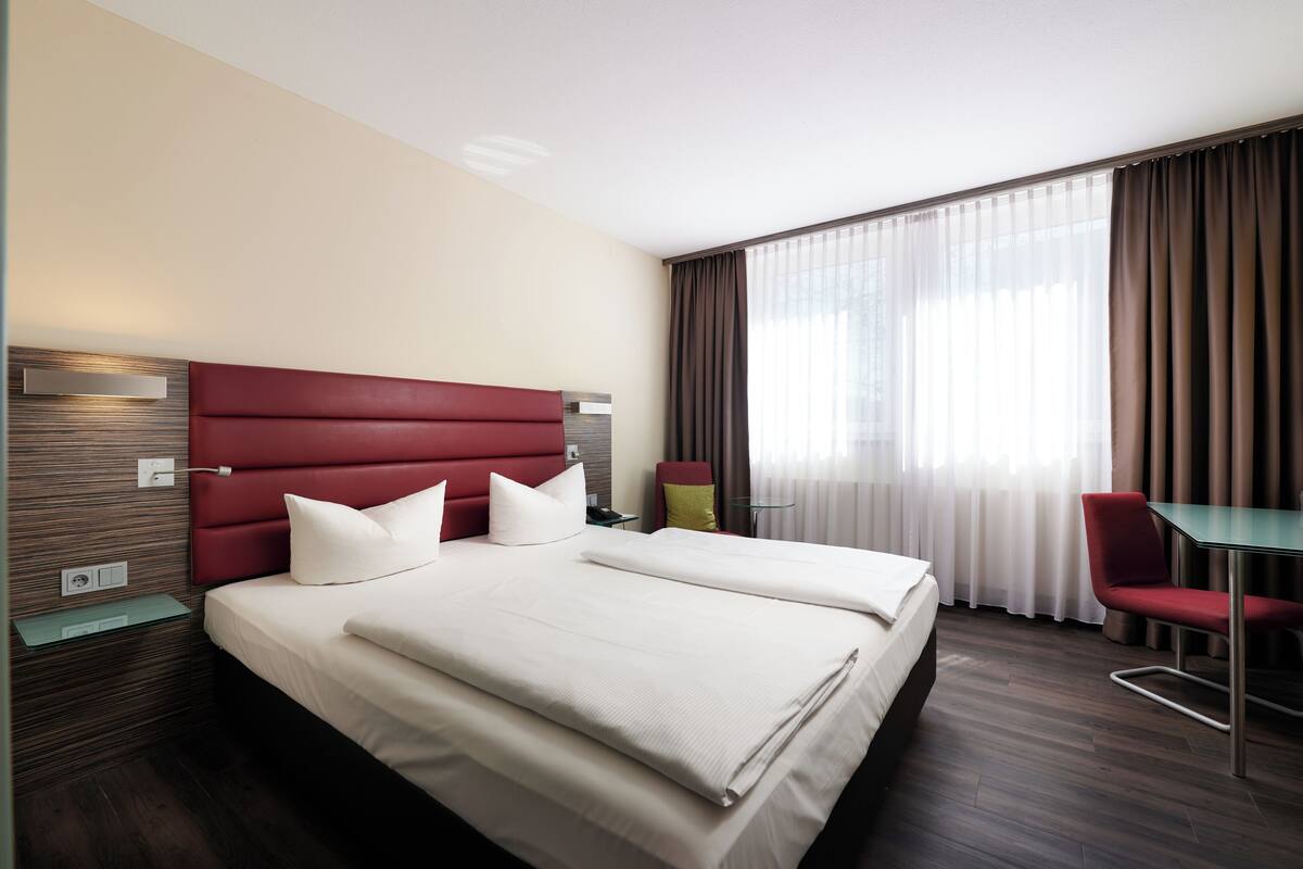 A hotel room with a large bed, a desk, a chair, and a window with curtains.