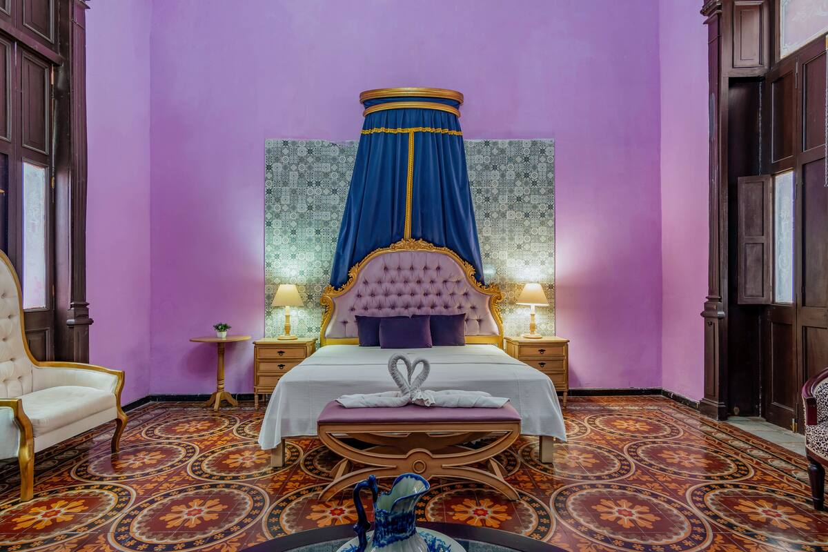 Quarto com cama grande, dossel, mesa de cabeceira, cadeira e um vaso decorativo.
