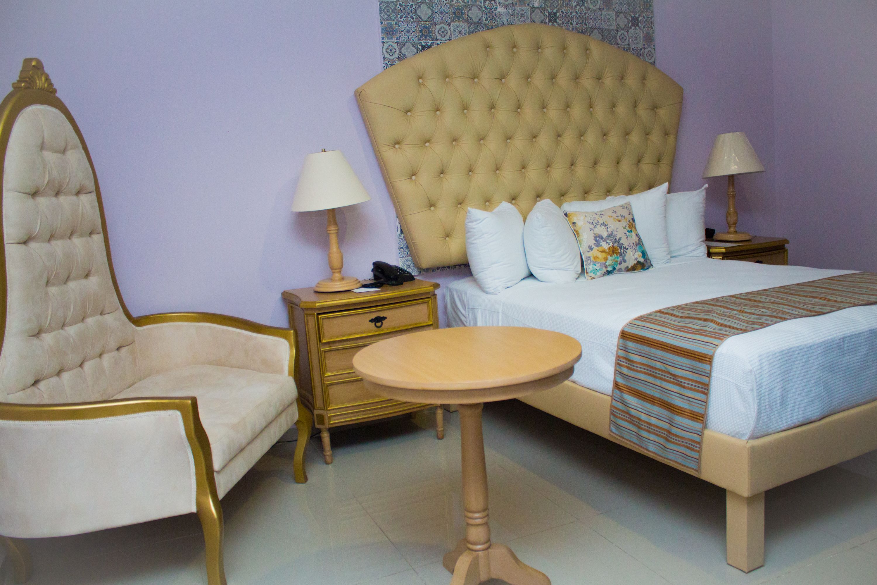 A bedroom with a large bed, a chair, a small table, and a nightstand with a lamp.