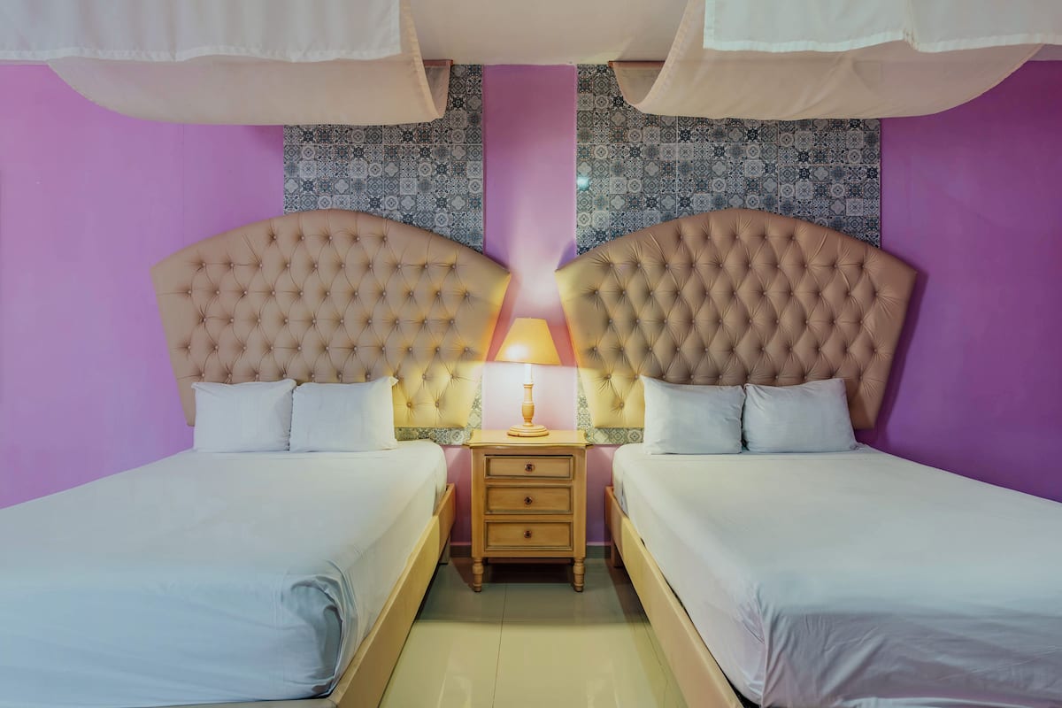 Quarto duplo com duas camas, uma mesa de cabeceira com abajur e paredes com padrão.