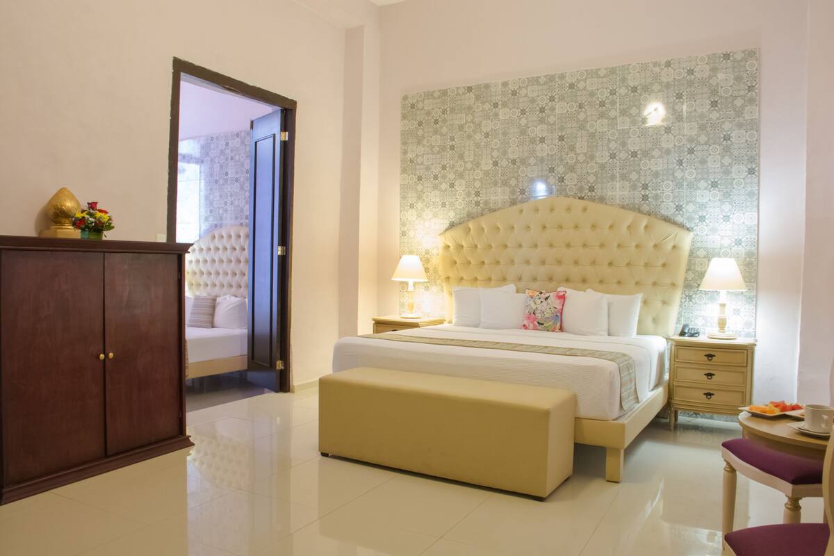 Quarto de hotel com uma cama grande, um guarda-roupa de madeira, uma mesa de cabeceira com abajur e uma pequena mesa redonda com uma cadeira.