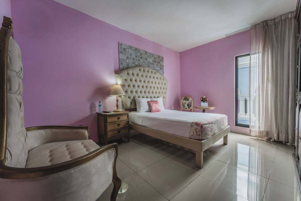 Quarto com uma cama grande, uma mesinha de cabeceira, uma cadeira e uma janela com cortinas.