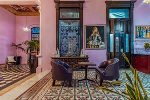 Un vestíbulo con piso a cuadros, una mesa central, dos sillones de cuero, un espejo decorativo y un cuadro grande en la pared.