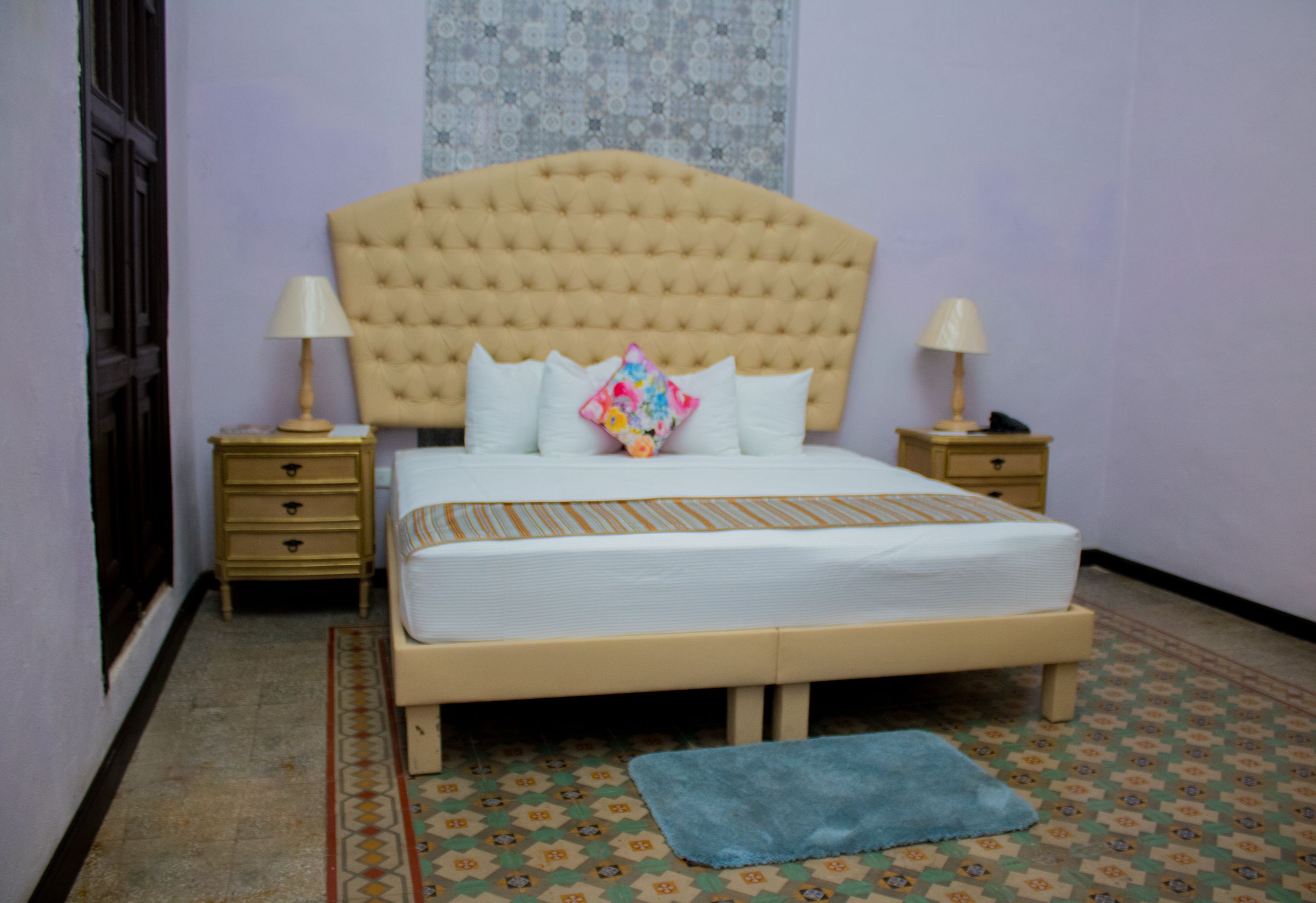 A bedroom with a tufted headboard, a bed with white bedding, two bedside tables with lamps, and a patterned floor.