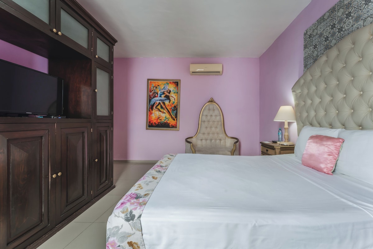 A bedroom with a bed, a television, a chair, a nightstand, and a painting on the wall.
