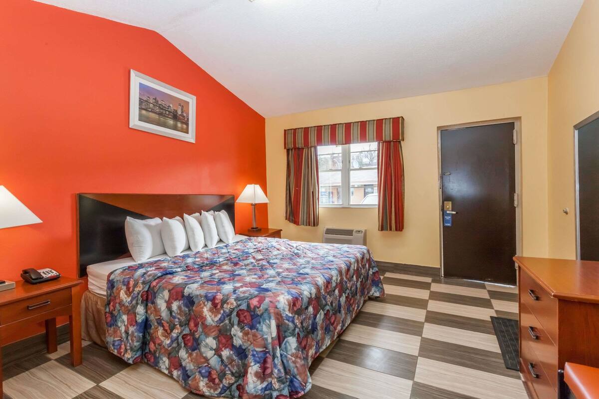 A hotel room with a large bed, a nightstand, a lamp, a window with curtains, a door, and a checkered floor.