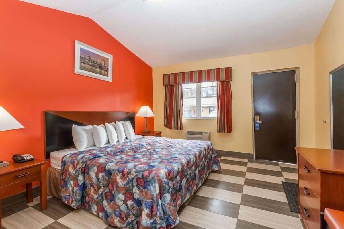 A hotel room with a bed, a nightstand, a lamp, a window with curtains, and a door.