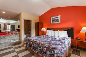 A hotel room with a large bed, two bedside lamps, a headboard, a painting on the wall, and a bathroom with a red sink and mirror.