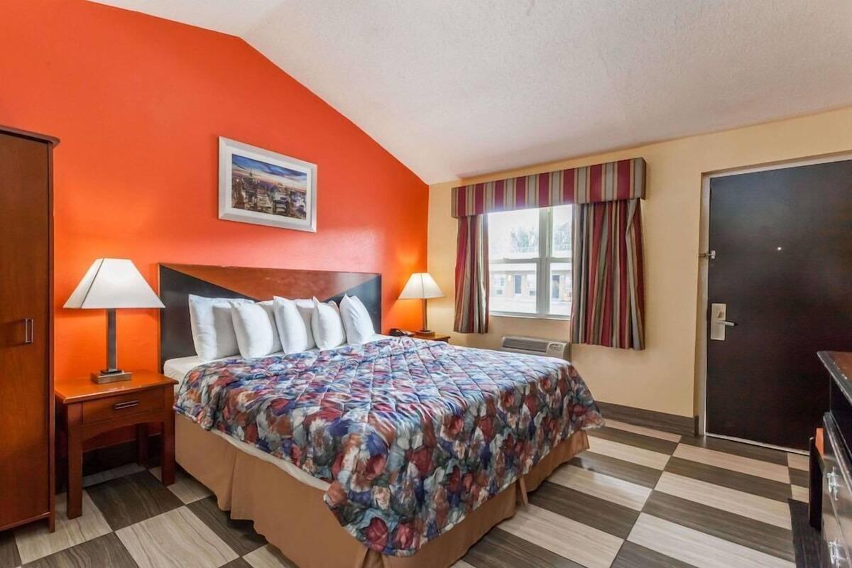 A hotel room with a bed, two bedside lamps, a window with curtains, a door, and a framed picture on the wall.