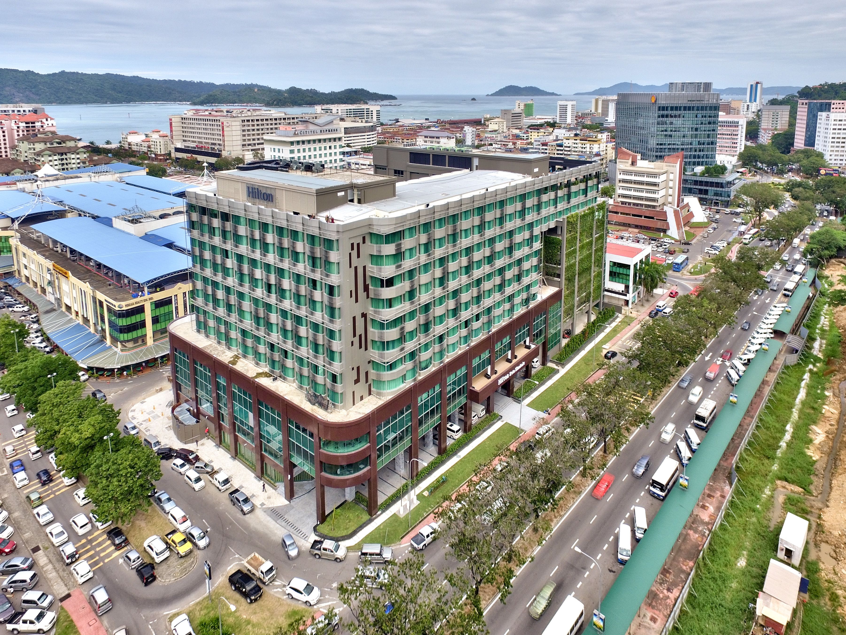 A multi-story hotel with a modern design, featuring a prominent Hilton sign, surrounded by a busy urban street with visible traffic and a waterfront view.