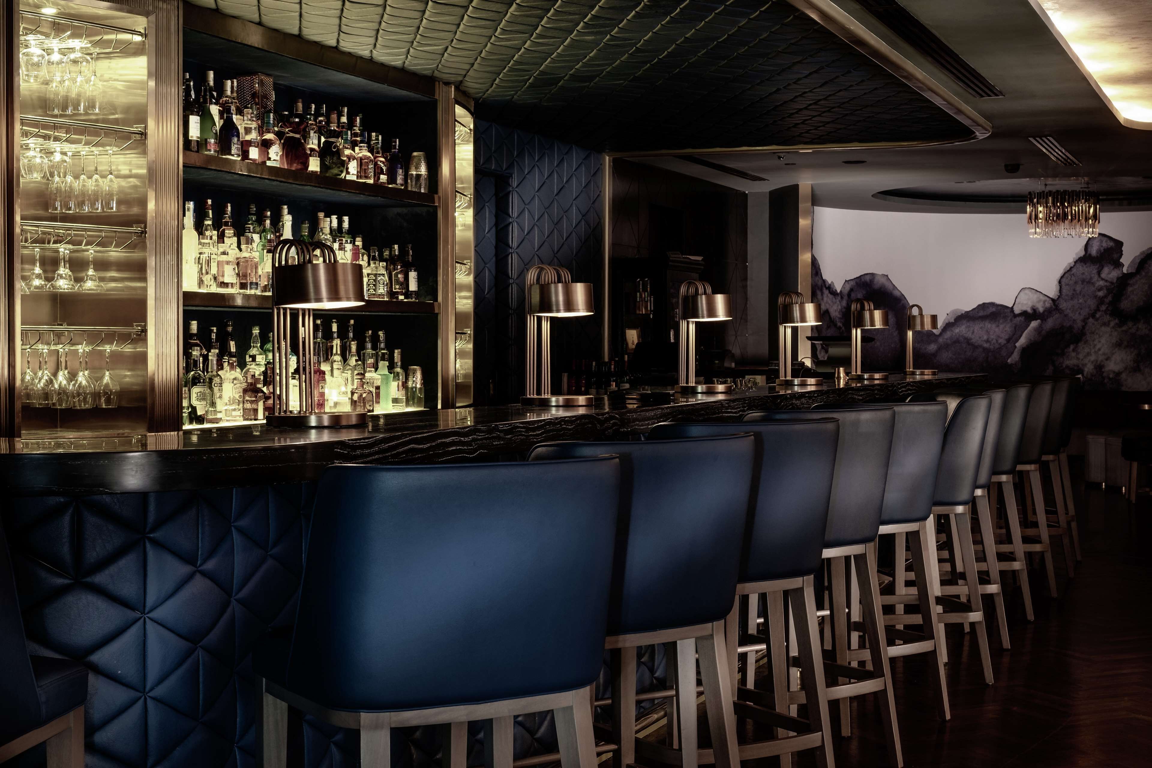 A bar area with a long counter, multiple shelves of bottles, and a row of blue bar stools.