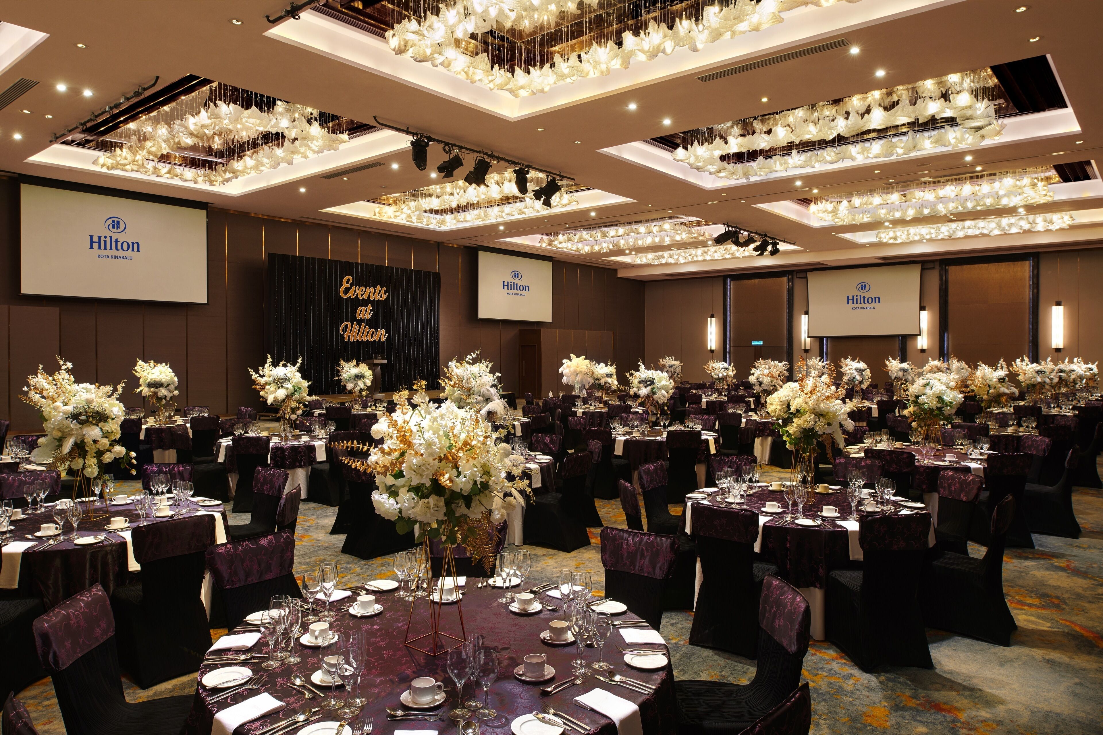 Une grande salle de banquet, bien éclairée, avec des tables rondes disposées pour les événements, agrémentée de compositions florales et d’une décoration de table élégante.