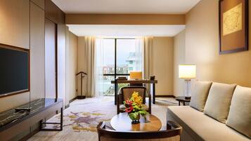 A hotel room with a flat-screen TV, a sofa, a dining table with a vase of flowers, and a view of the city through large windows.