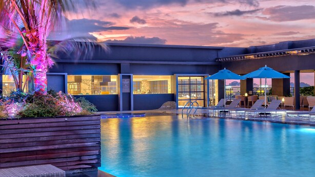 A swimming pool with lounge chairs and umbrellas, surrounded by a modern building with large windows. - Hilton Kota Kinabalu (Kota Kinabalu)