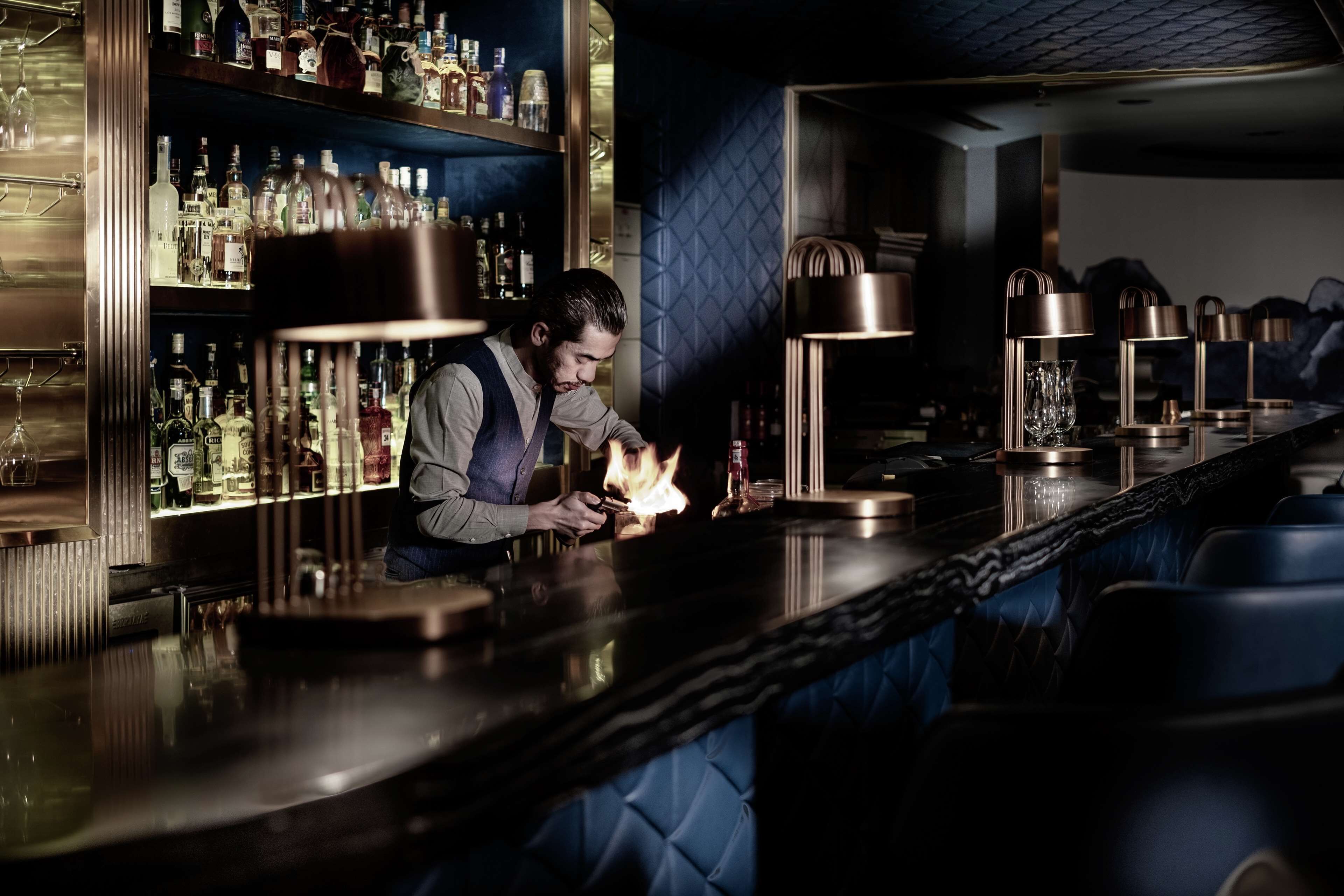 A bar area with a bartender preparing drinks, shelves with bottles, and a blue cushioned seating area.