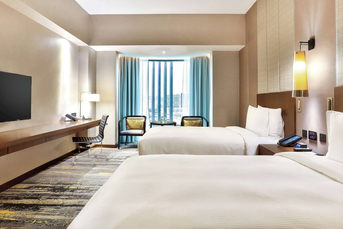 Ein Hotelzimmer mit zwei Betten, einem Fernseher, einem Schreibtisch und Blick auf die Stadt durch große Fenster.