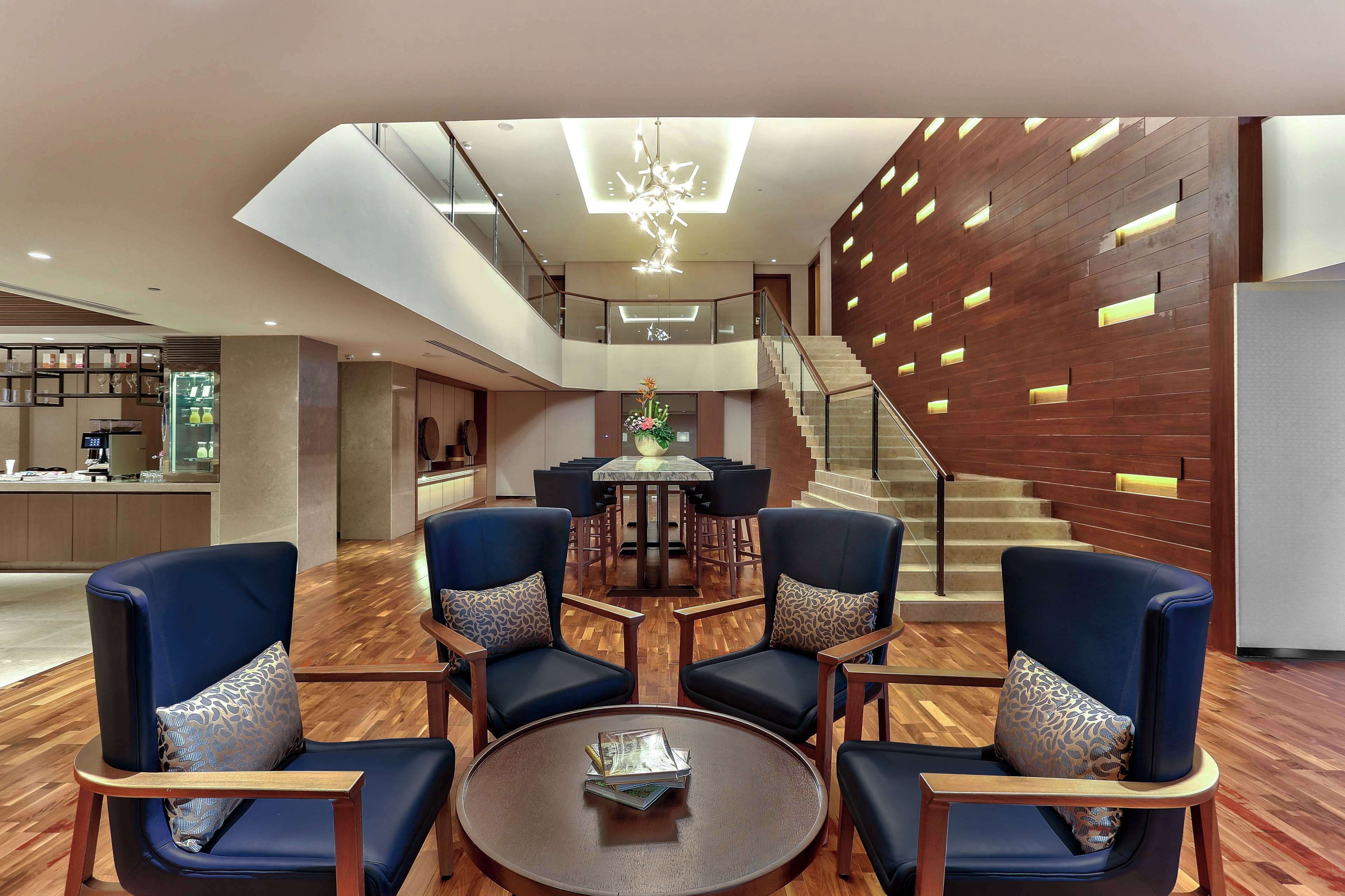 A spacious lobby with a central round table surrounded by blue armchairs, a staircase with wooden railings, and a modern ceiling light fixture.