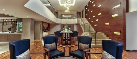 A spacious lobby with a central round table surrounded by blue armchairs, a staircase with wooden railings, and a modern ceiling light fixture.