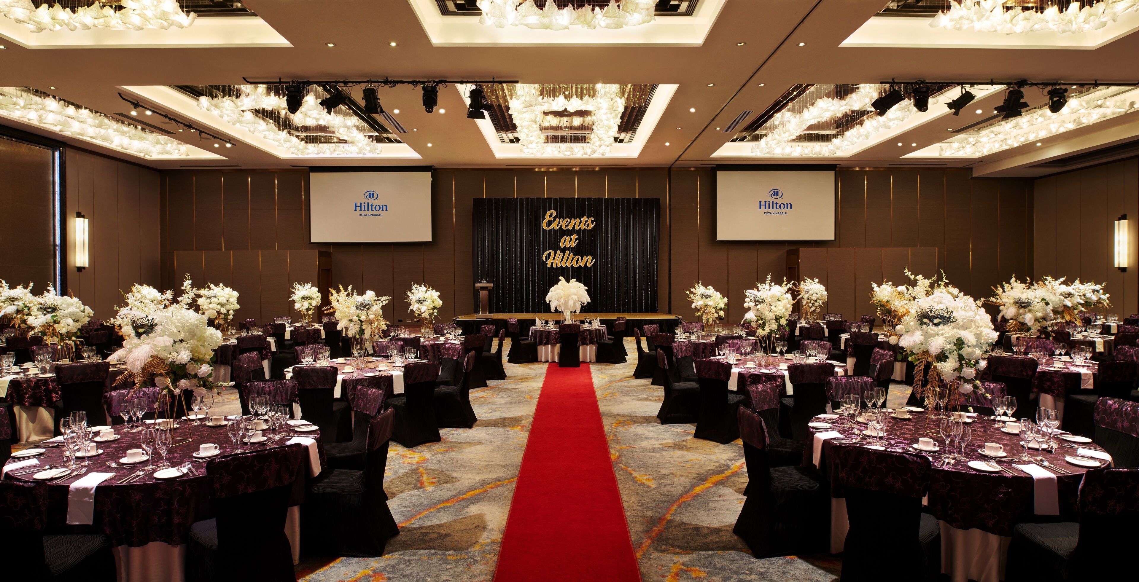 A banquet hall with round tables, red carpet, and floral centerpieces.