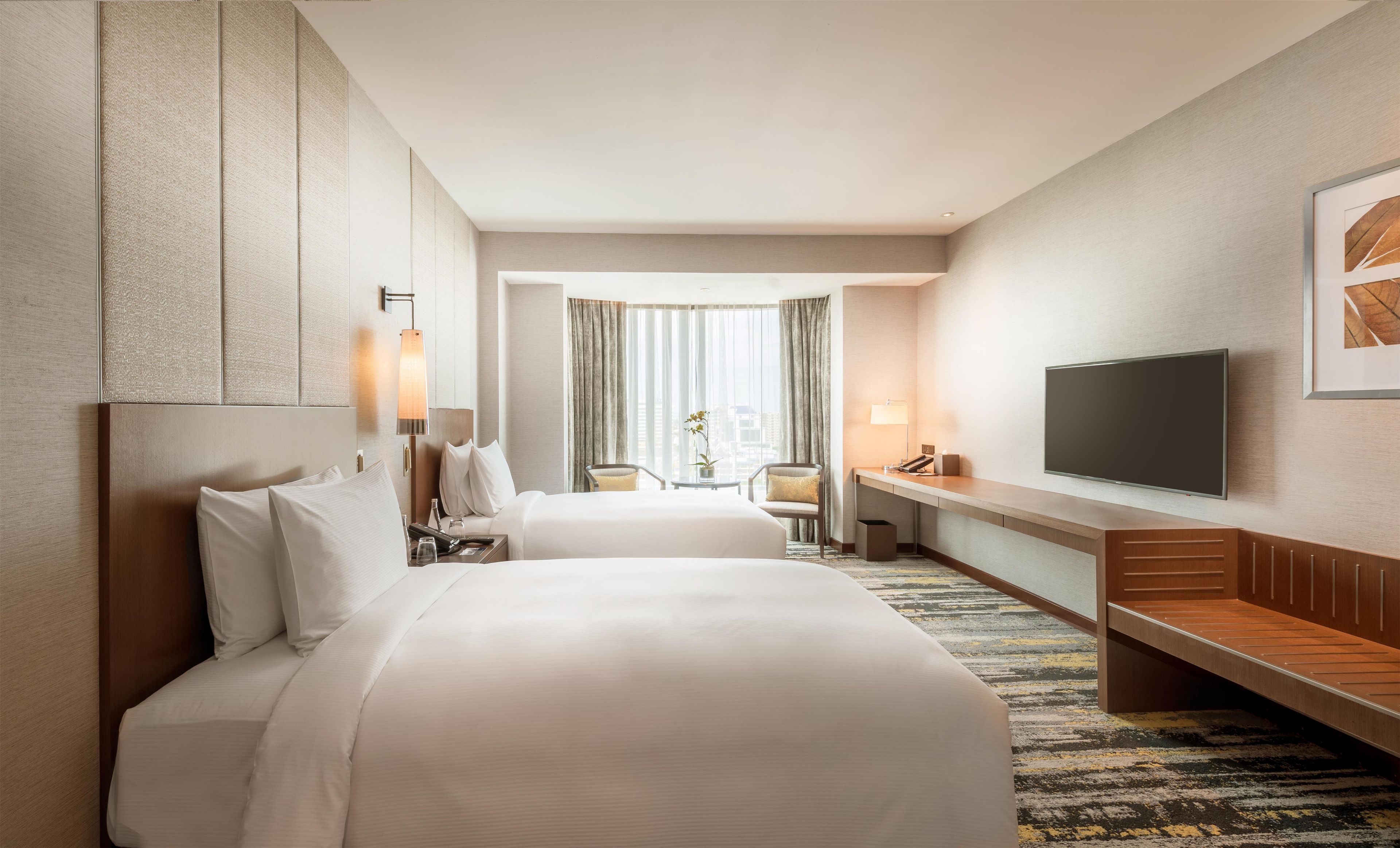 A hotel room with a large bed, a desk with a flat-screen TV, a chair, and a small table with a vase of flowers.