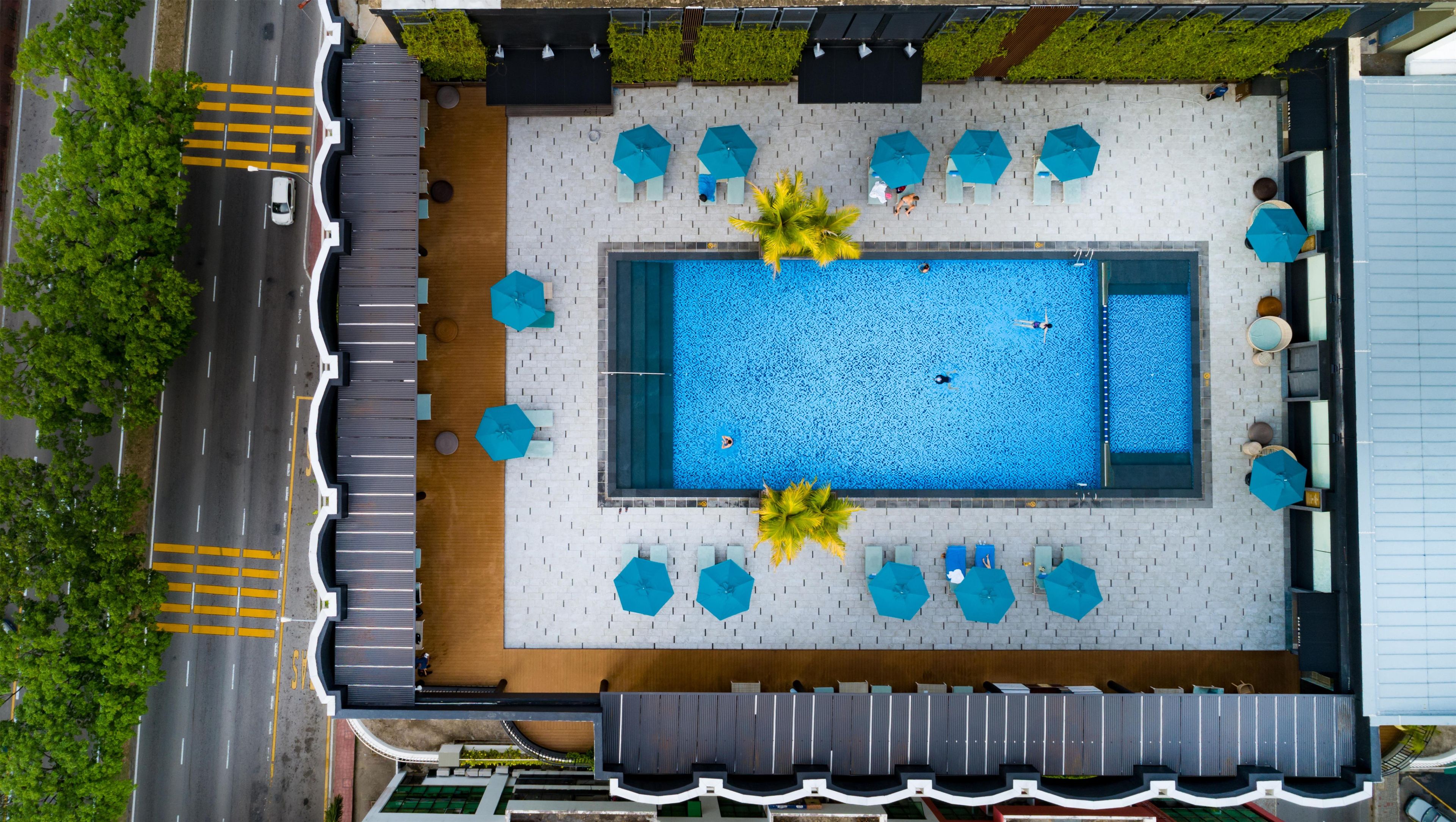 An aerial view of a rooftop pool area with a rectangular pool, surrounded by lounge chairs and umbrellas, and a road adjacent to the pool.