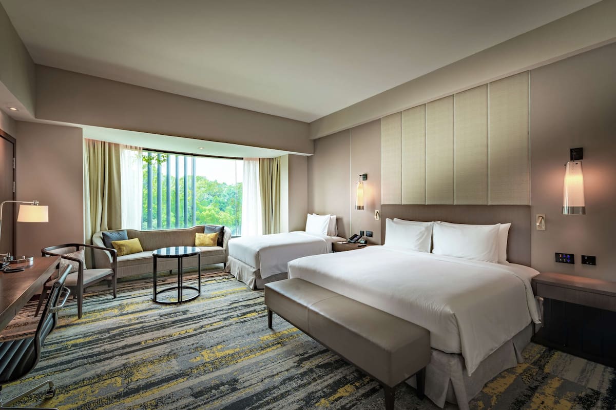 Ein Hotelzimmer mit zwei Betten, einer Couch, einem Schreibtisch und Blick auf Grünflächen.