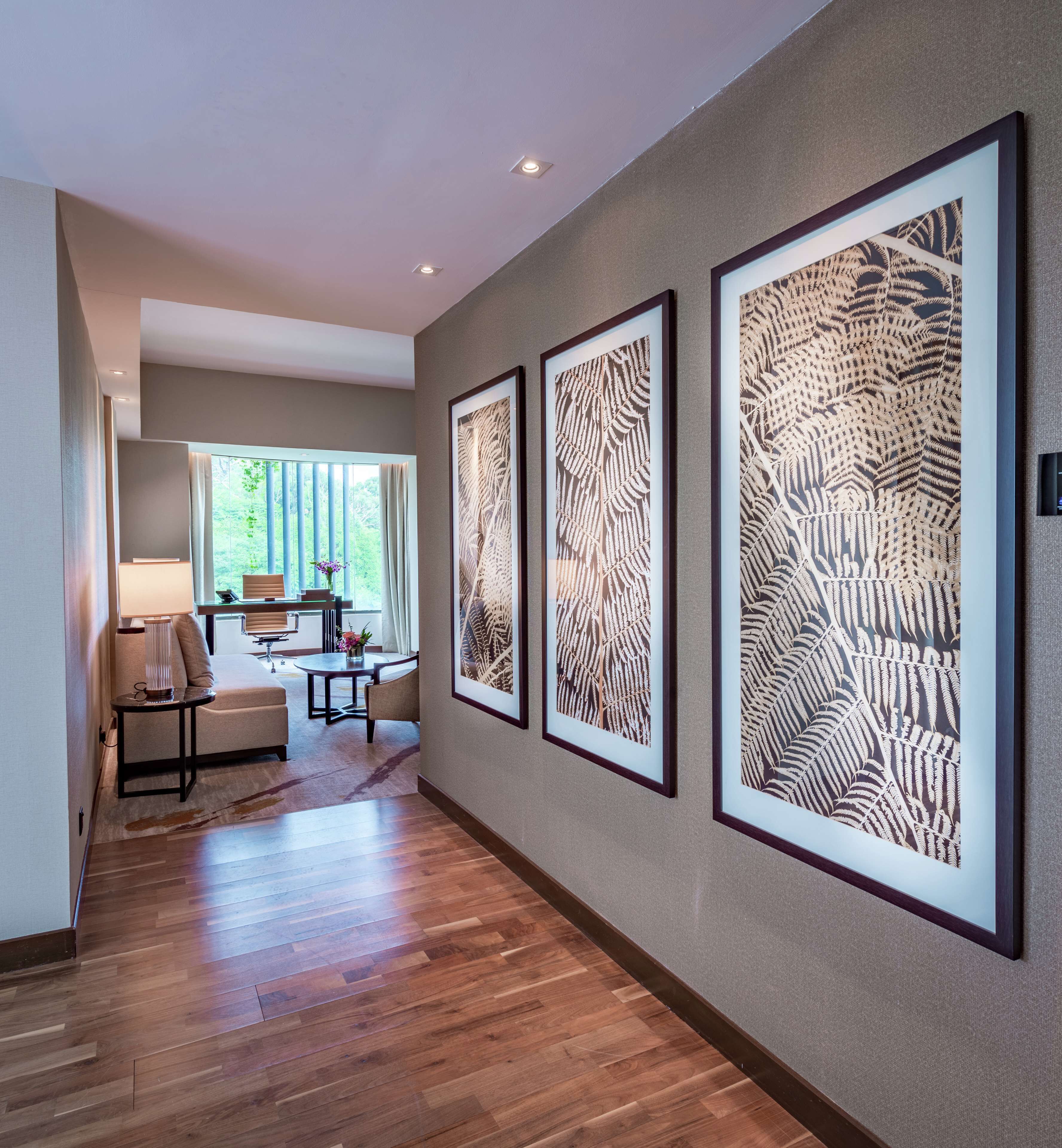A hallway with wooden flooring, framed artwork on the wall, a seating area with a table and chairs, and a window with curtains.
