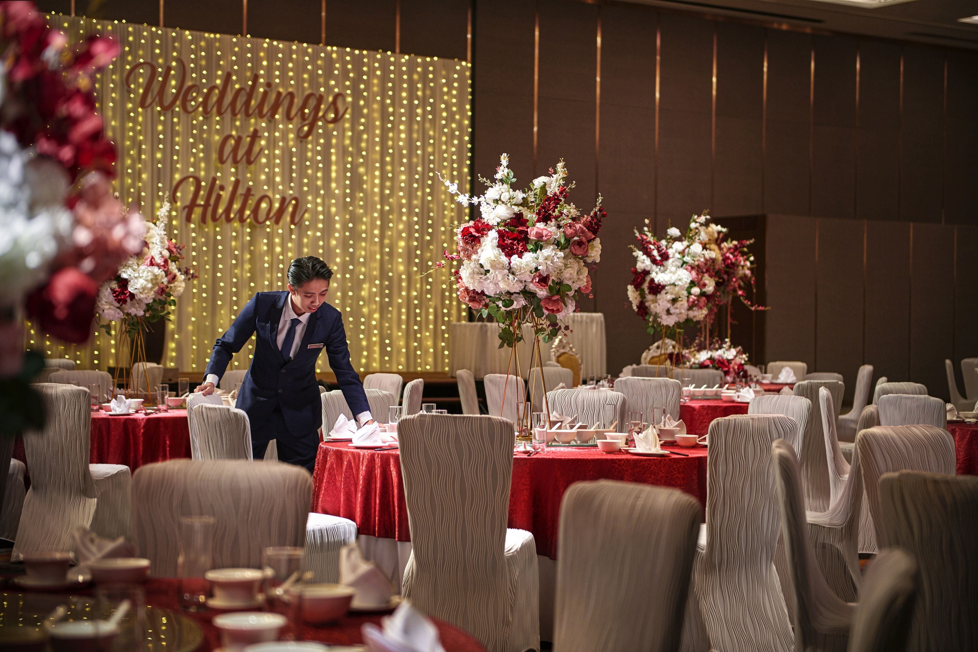 A banquet hall with tables set for a wedding, featuring red tablecloths and floral centerpieces.