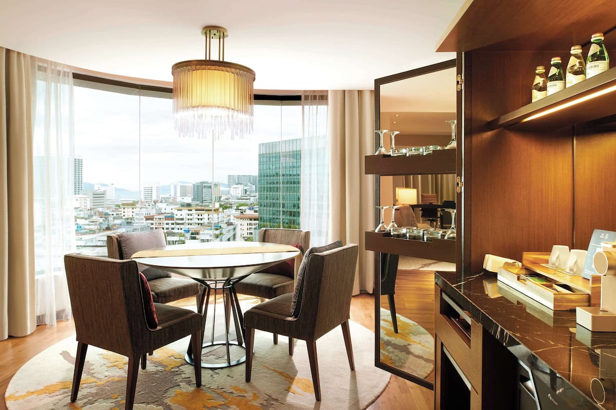 Ein Hotelzimmer mit einem runden Esstisch, vier Polsterstühlen, einer Chandelieber, einem großen Fenster mit Stadtblick und einer Minibar mit verschiedenen Artikeln.