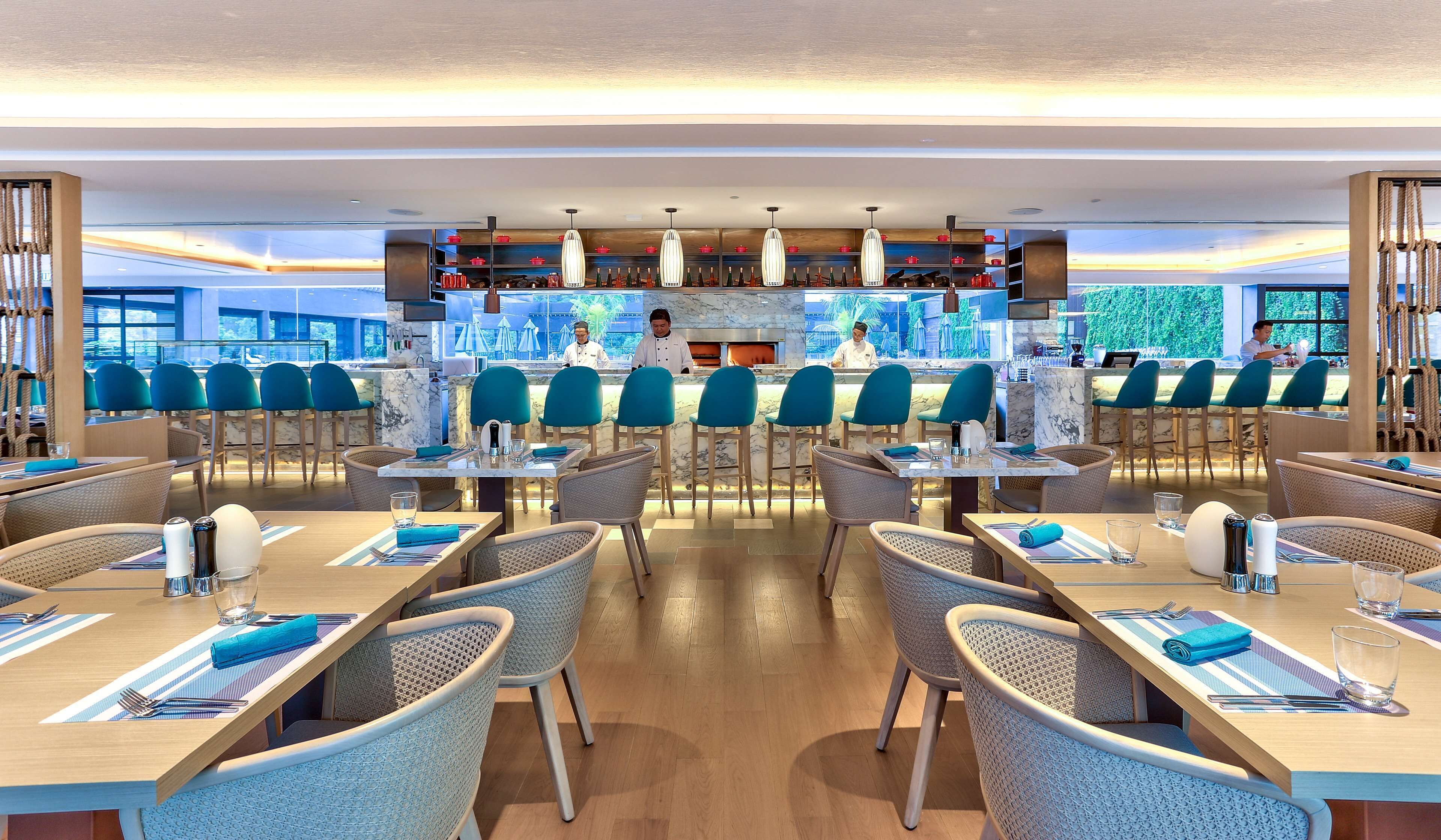 A modern restaurant with a central bar area, multiple seating arrangements, and a view of the outdoors.
