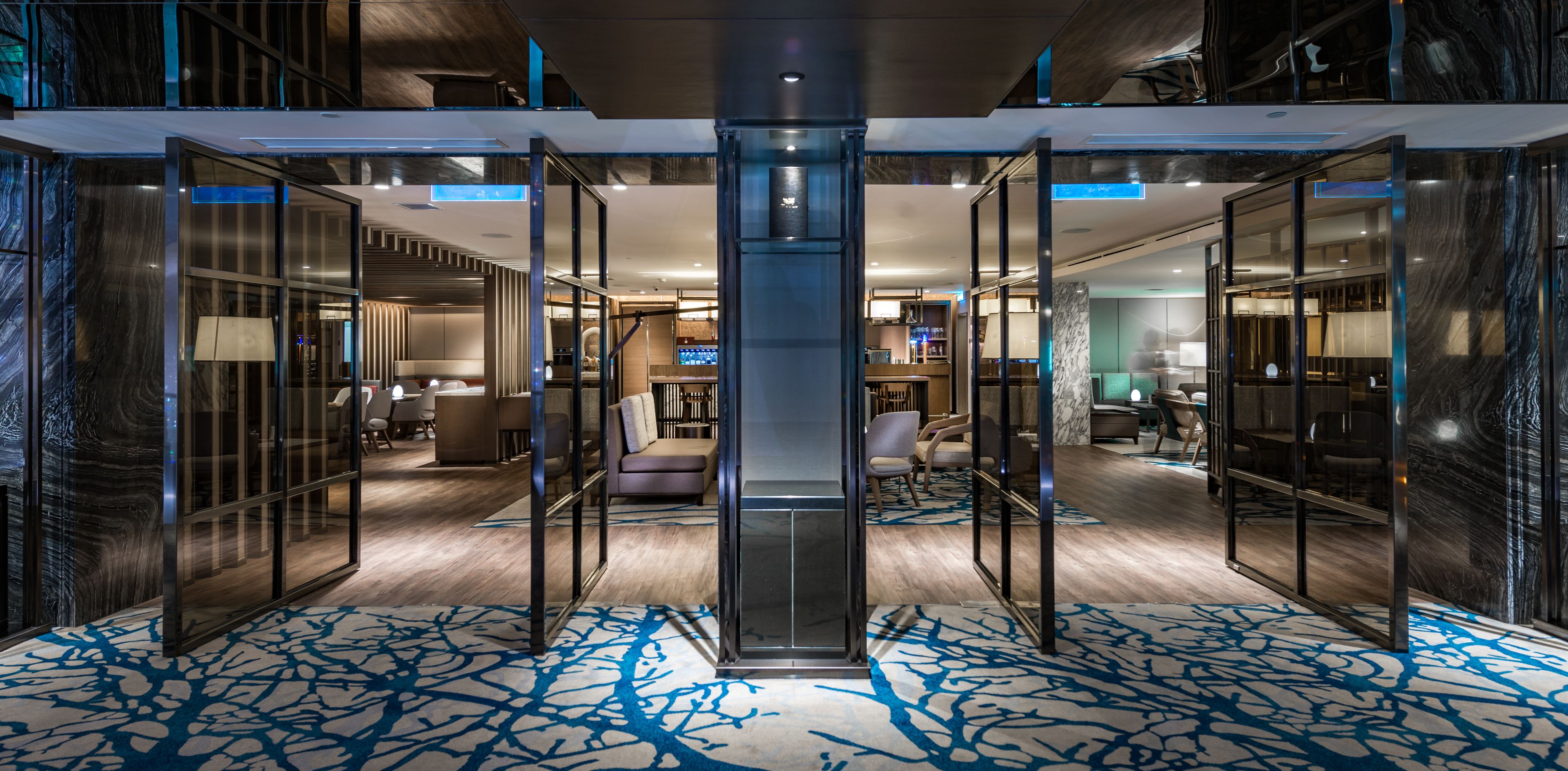 A modern hotel lobby with a central elevator, glass partitions, and a patterned carpet.