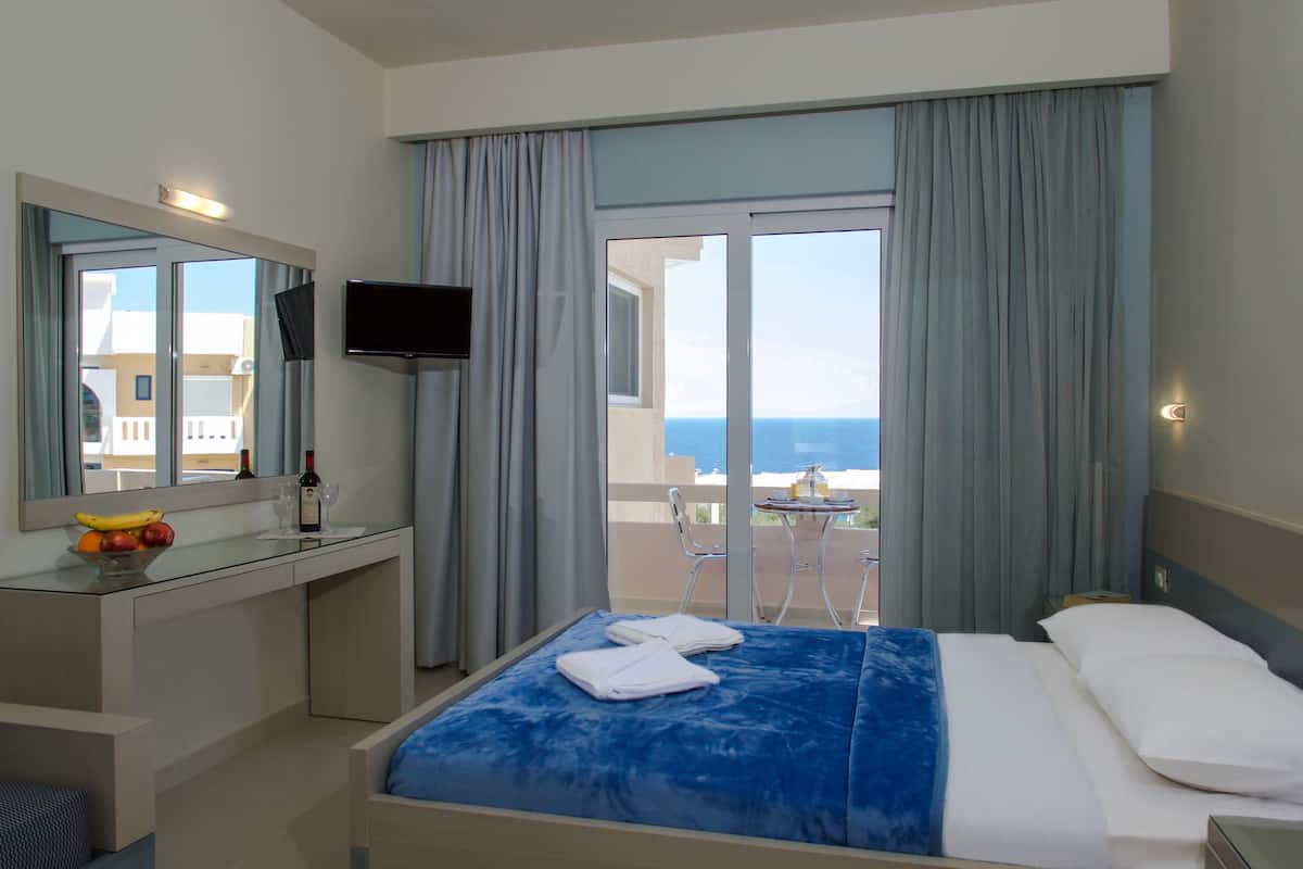 Habitación de hotel con cama, escritorio con televisor, frutero y vista al mar.
