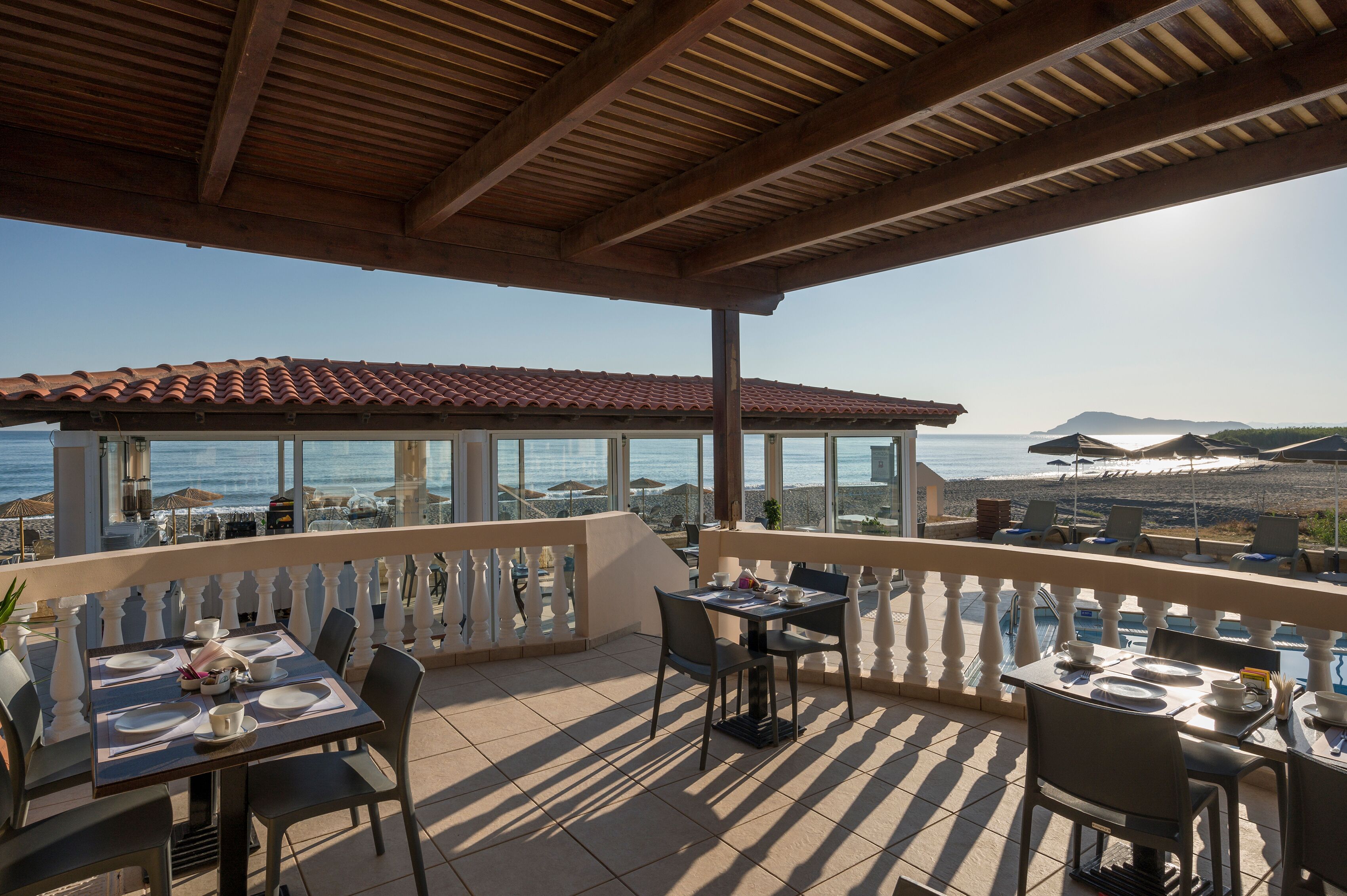 Eine Terrasse mit Esstischen, mit Blick auf einen Strand und das Meer.