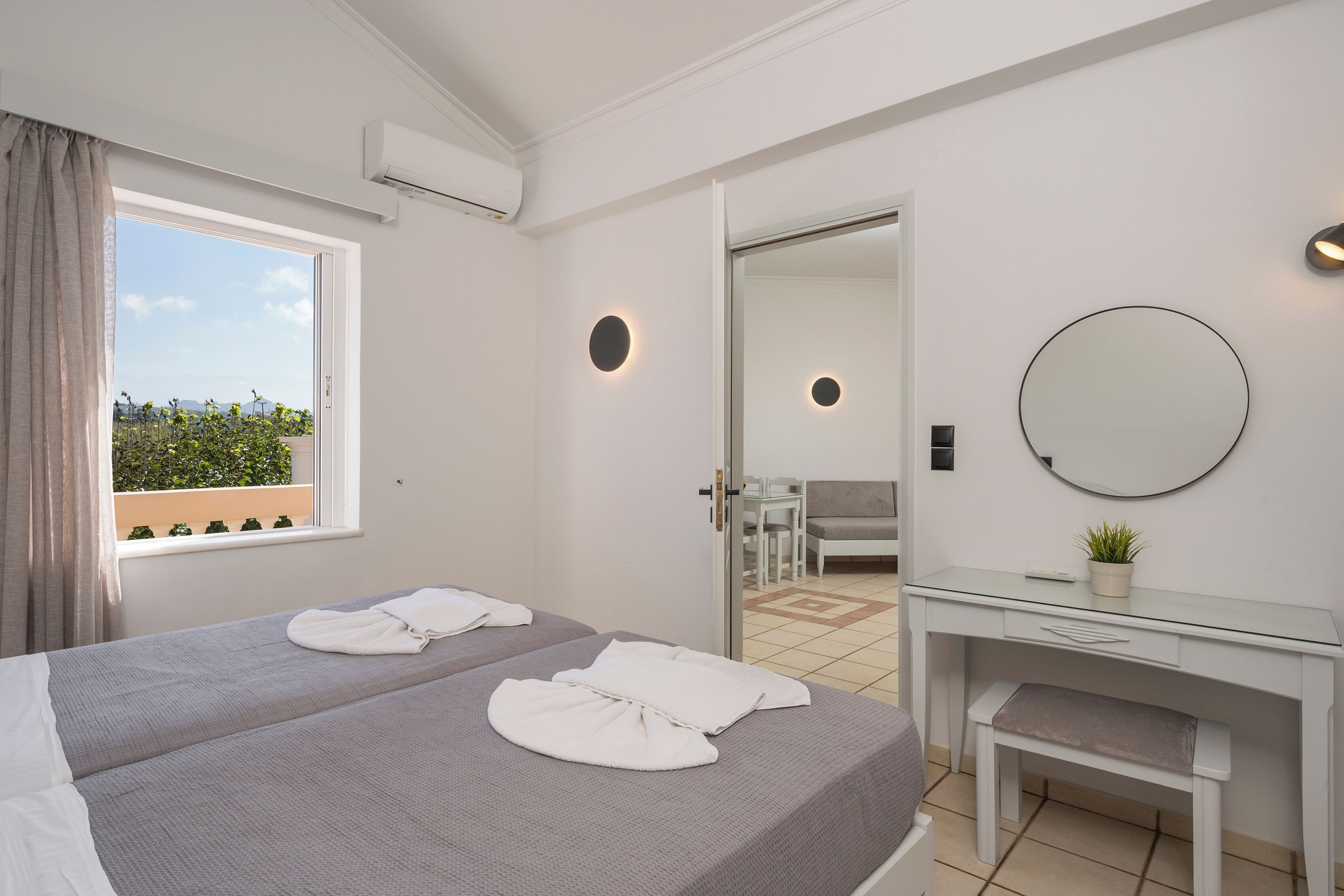 A hotel room with two beds, a desk, a mirror, and a view of the outdoors.