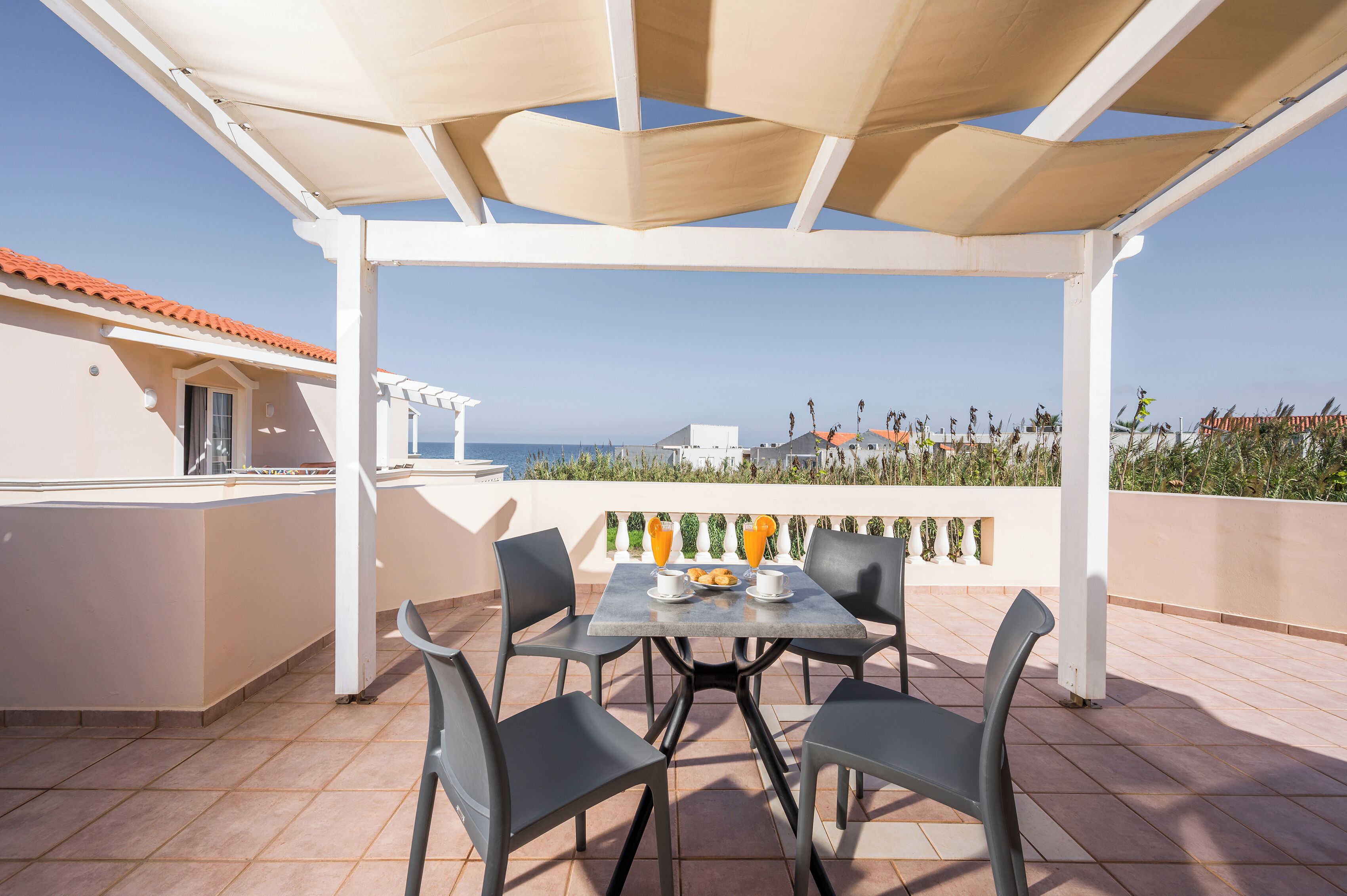 A terrace with a table and chairs, a view of the sea, and a building with a red-tiled roof.