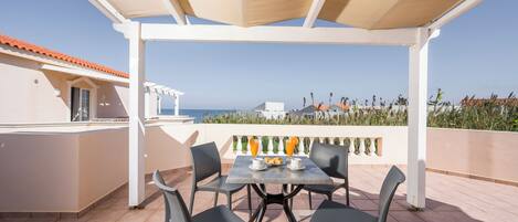 A terrace with a table and chairs, a view of the sea, and a building with a red-tiled roof.