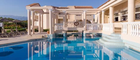 A swimming pool with a clear blue water, surrounded by white columns and a covered area with a roof.