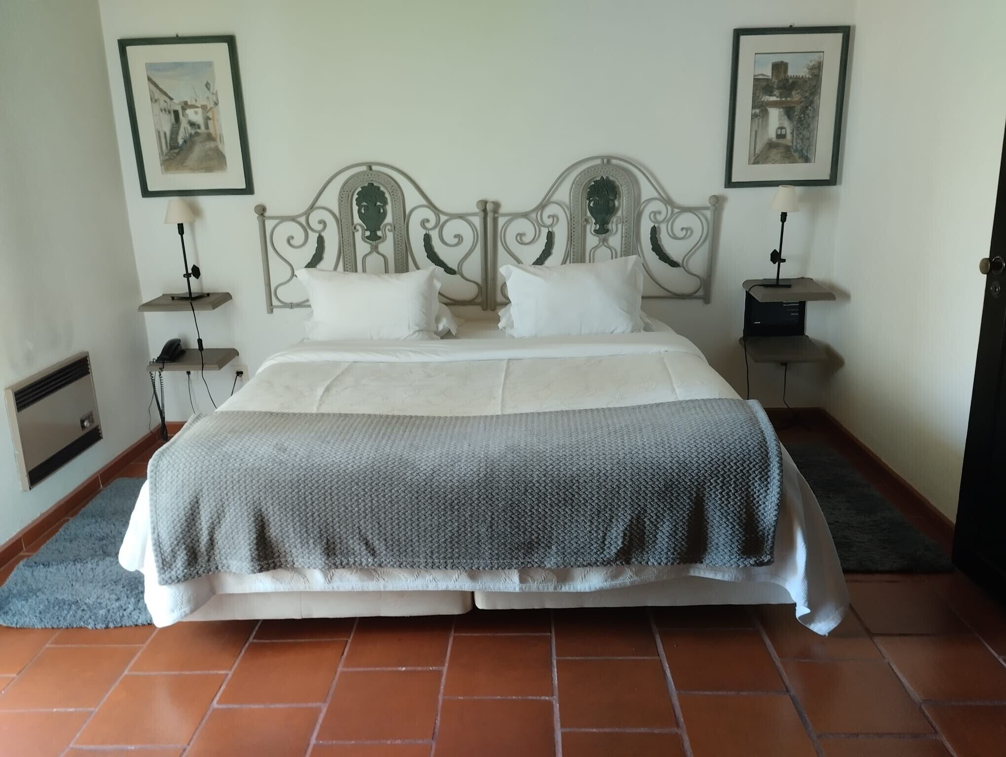 A neatly made bed with a grey and white duvet, two white pillows, and a decorative metal headboard. There are two bedside tables with lamps, and two framed pictures on the walls.