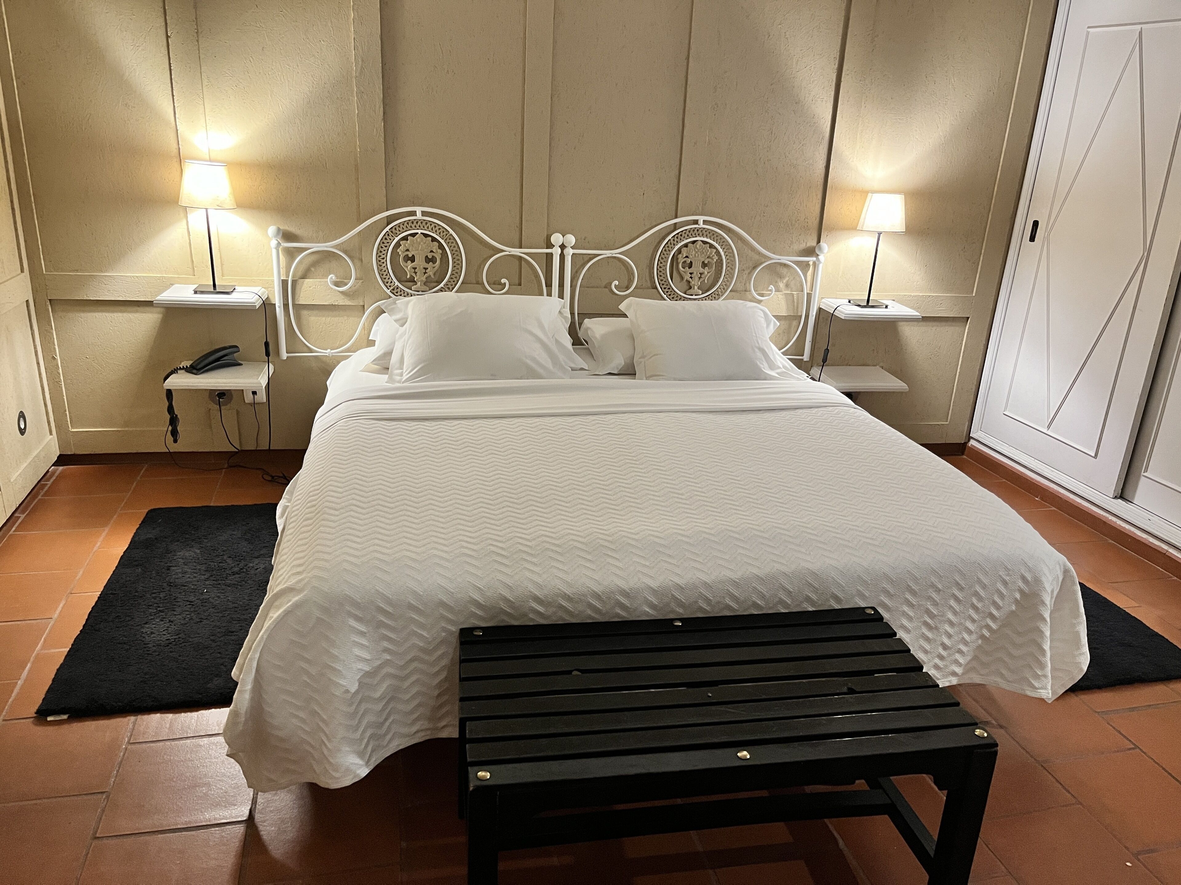 A neatly made bed with white linens, two bedside tables with lamps, a black bench, and a tiled floor.